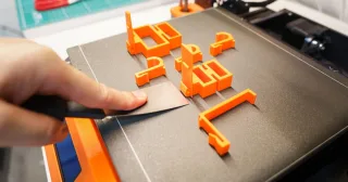 Mão retirando peças laranja impressas em 3D da mesa com espátula (foto ilustrativa)