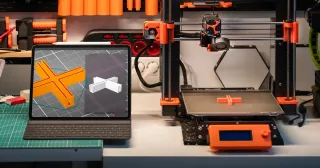 Tablet com modelo 3D ao lado de uma impressora 3D imprimindo a peça (foto ilustrativa)
