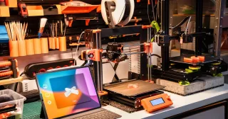 Bancada de oficina com impressoras 3D, tablet e ferramentas (foto ilustrativa)